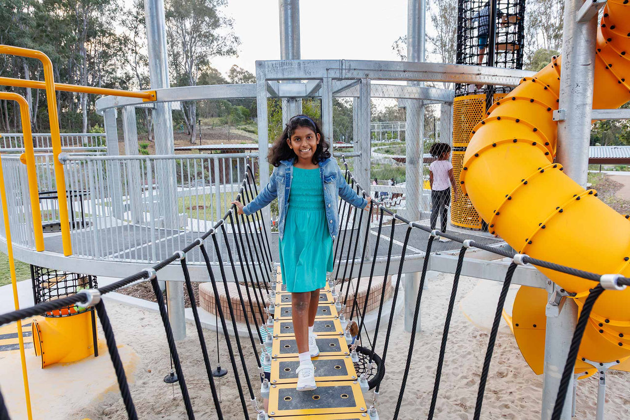Girl on rope bridge with metal platform at Archerfield Wetlands District Park playground.