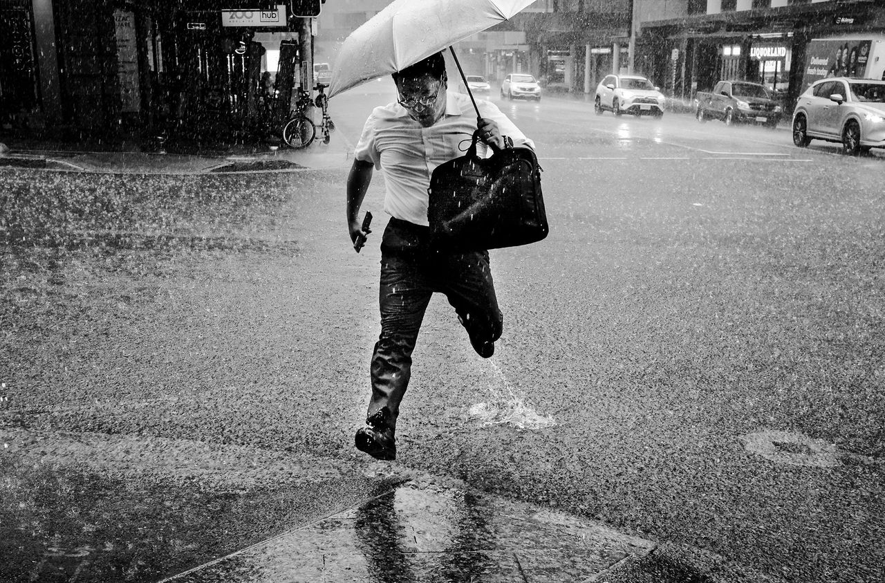 ord Mayor's Photographic Awards 2026 - Open Category Winner - Tyler Sharman - Rush hour. Black and white image of man holding an umbrella running across a city street during rain.