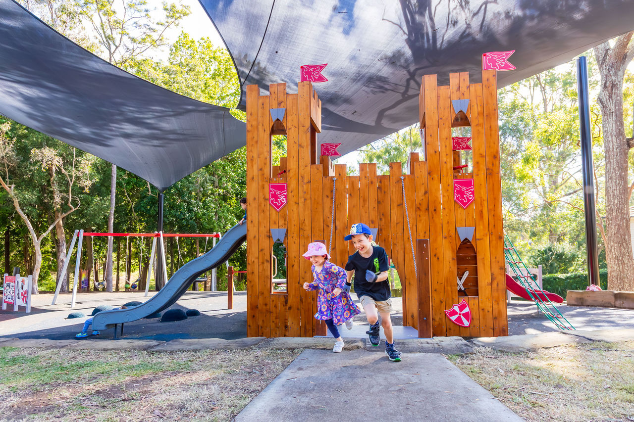 2 kids running in Majestic Park, Coorparoo playground. The area is covered by dark blue shade sails.